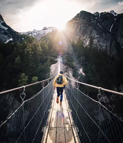Hiker crossing a suspension bridge towards sunlit mountains